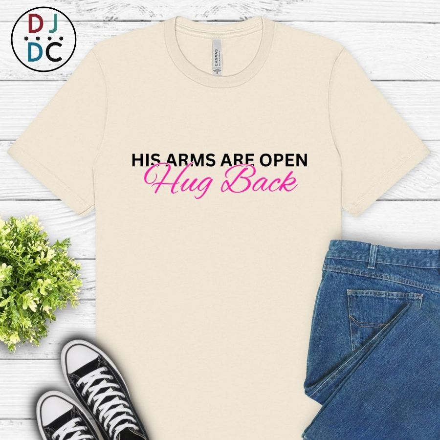 Beige t-shirt with text 'His arms are open Hug Back' on a wooden surface with jeans and sneakers.