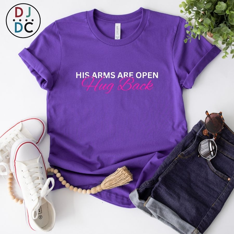 Purple t-shirt with text 'His arms are open, Hug Back' on a white surface with accessories.